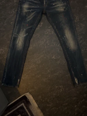 Dsquared2 jeans - Säljer ett par mörkblå jeans med coola slitningar och vita färgstänk. Jeansen har klassisk femficksmodell, knappgylf och smal passform. Perfekta för dig som gillar en trendig och avslappnad stil.