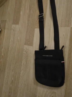 Svart axelväska Tommy Hilfiger - Snygg svart axelväska från Tommy Hilfiger med justerbar axelrem och diskret logga framtill. Väskan har en enkel, rektangulär form och en ficka på framsidan. Materialet ser ut att vara slitstark textil, perfekt för att bära det viktigaste.