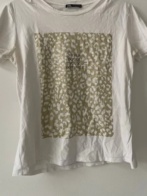 Vit t-shirt med leopardmönster Zara - Vit t-shirt från Zara med beige och vitt leopardmönster framtill och texten 'Never doubt your instinct' tryckt i svart. Klassisk rund hals och korta ärmar. Perfekt för dig som vill ha en trendig och cool look.