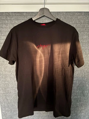 Svart t-shirt med röd Hugo-logga - Snygg svart t-shirt från Hugo med röd broderad logga på bröstet. Klassisk passform och rund halsringning. Perfekt basplagg som funkar till det mesta. Tillverkad i mjuk bomull för skön känsla hela dagen.