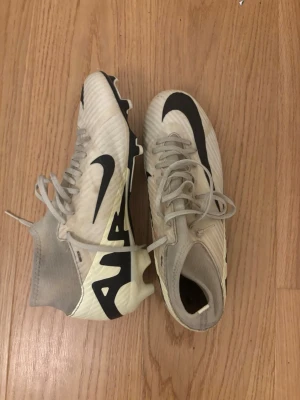 Vita Nike fotbollsskor med högt skaft - Säljer ett par vita Nike fotbollsskor med högt skaft och svarta detaljer. Skorna har snörning, en stor svart Nike-logga på sidan och mönstrad sula för bra grepp på planen. Materialet är syntetiskt och designen är modern och sportig.