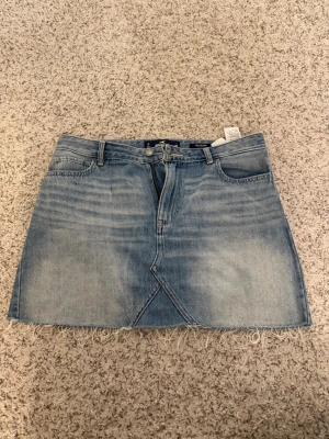 Ljusblå jeanskjol med fransig kant från hollister - Säljer en ljusblå jeanskjol i kort modell med fransig rå kant nedtill. Kjolen har klassiska fem fickor, bälteshällor och en snygg asymmetrisk söm framtill. Perfekt till en avslappnad och trendig look.