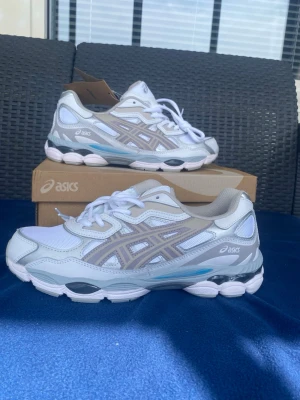 Asics skor  - Snygga sneakers från Asics i vitt och ljusgrått med mesh och syntetpaneler. Skorna har klassisk Asics-logga på sidan, vita skosnören och detaljer i ljusblått på sulan. Perfekta för dig som gillar sportig och trendig stil. Boxen kommer ej med endast skorna 