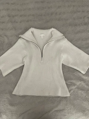 Vit ribbstickad tröja - Vit ribbstickad tröja med halv dragkedja och bred krage. Tröjan har trekvartsärmar och är tillverkad i ett mjukt, stretchigt material. Perfekt för lager-på-lager och en clean look.