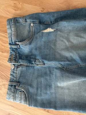 Ljusblå slitna jeans med hög midja - Säljer ett par ljusblå jeans med hög midja och en snygg sliten detalj på låret. Klassisk femficksmodell med dragkedja och knapp. Perfekta för en avslappnad och trendig look.
