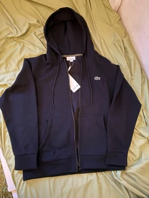 Svart hoodie med dragkedja från Lacoste - Snygg svart hoodie från Lacoste med dragkedja framtill och klassisk krokodillogo på bröstet. Hoodien har huva med dragsko och två stora fickor framtill. Tillverkad i mjuk bomull för skön känsla och stilren look.