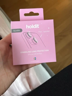 Holdit sparkling lens protection silver - Snyggt glittrande linsskydd i silver från Holdit, designat för iPhone 14 och 14 Plus. Skyddet ger extra stil och skydd till kameran på din mobil. Förpackningen är rosa och visar hur skyddet ser ut på telefonen. Kartongen är öppnad men själva produkten är orörd 
