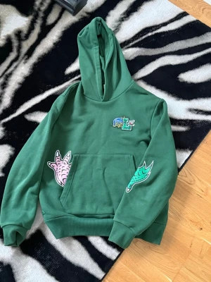 Grön Lacoste Hoodie - Grön hoodie från Lacoste med stor huva och känguruficka. Dekorerad med färgglada tryck, bland annat en rosa klo och gröna detaljer. Tillverkad i mjukt bomullsmaterial och har ribbade muddar vid ärmslut och nederkant.