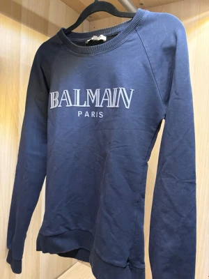 Mörkblå sweatshirt från Balmain - Snygg mörkblå sweatshirt från Balmain Paris med tryckt logga på bröstet. Tröjan har rund halsringning, långa ärmar och ribbade muddar vid ärmslut och nederkant. Tillverkad i mjukt bomullsmaterial för en bekväm känsla.