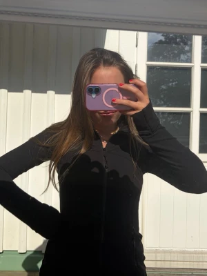 Svart långärmad jacka med dragkedja - Säljer en svart, figurnära jacka med lång ärm och hel dragkedja framtill. Jackan har en enkel och stilren design som passar perfekt till vardags. Materialet ser ut att vara mjukt och stretchigt, vilket gör den bekväm att bära.