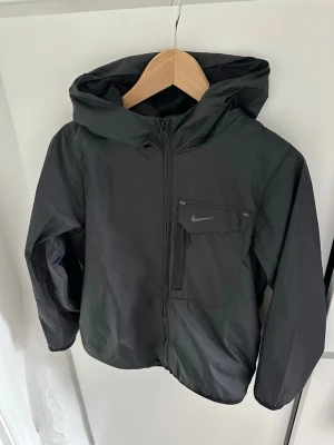 Svart vindjacka från Nike - Svart vindjacka från Nike med huva och dragkedja framtill. Jackan har en bröstficka med dragkedja och Nike-logga, samt en sportig och stilren design. Perfekt för blåsiga dagar och enkel att matcha med olika outfits. Storleken är 147-158 men passar som xxs