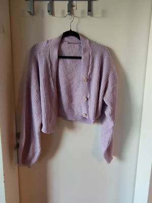 Ljuslila cropped stickad kofta - Säljer en ljuslila stickad kofta i croppad modell med tre knappar framtill. Koftan har bred ribbad krage och långa, lite pösiga ärmar. Perfekt för att slänga över en topp när det är lite kyligt. Materialet känns mjukt och mysigt.