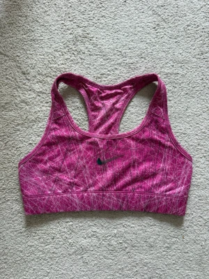 Rosa sport-bh från Nike med mönster - Snygg rosa sport-bh från Nike med vitt och svart grafiskt mönster. Modellen har brottarrygg och är tillverkad i ett stretchigt funktionsmaterial som passar perfekt för träning. Nike-logga framtill som blivit lite sliten. 