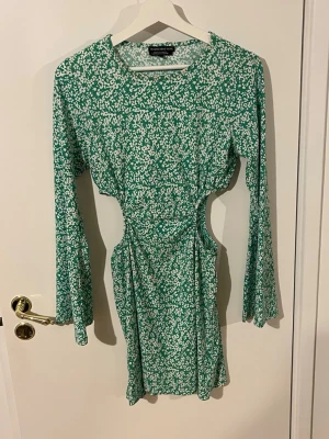 Grön blommig cut out klänning - Grön kort klänning med vitt blommönster och långa ärmar. Klänningen har trendiga cut out-detaljer i midjan och en figurnära passform. Perfekt för dig som vill sticka ut med en cool och modern look. Enbart andvänd 1 gång, Storlek S men passar även XS och M. Pris går att diskutera vid snabb affär.