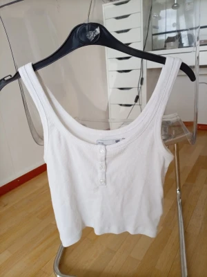 Vit ribbad linnetopp med knappar - Vit ribbad linnetopp från 157 med rundad halsringning och tre dekorativa knappar framtill. Toppen är ärmlös och har en croppad passform som passar perfekt till jeans eller kjol.