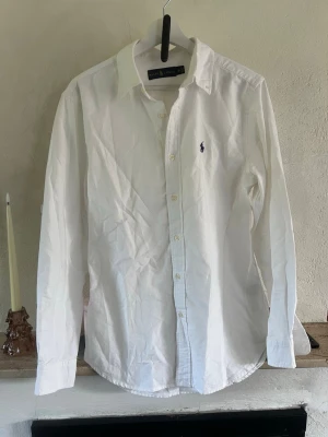 Vit skjorta från Ralph Lauren - Klassisk vit skjorta från Ralph Lauren med blå broderad logga på bröstet. Skjortan har lång ärm, knappar framtill och en tidlös krage. Perfekt till jeans eller chinos för en clean och stilren look.
