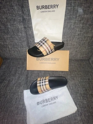  sandaler med rutigt band - Snygga sandaler   med klassiskt beige, svart och rött rutigt band över foten. Sulan är svart och har en bekväm form. Perfekta för sommaren och ger en lyxig touch till din outfit. Kommer med originalpåse och låda.