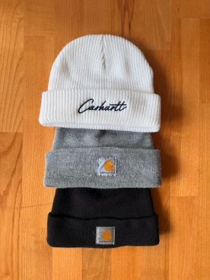 Tre Carhartt mössor i olika färger - Säljer tre snygga Carhartt mössor i ribbstickad modell. En vit med broderad logga, en grå och en svart med klassisk Carhartt-märke framtill. Perfekta för kalla dagar och ger en clean streetwear-look. Alla tre för 600kr eller 250kr st.