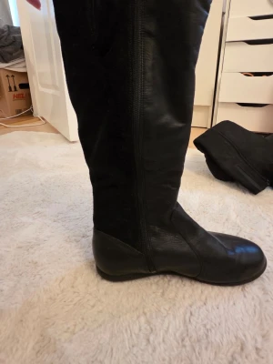 Svarta knähöga läderstövlar - Säljer ett par svarta knähöga stövlar i läder med dragkedja på sidan. Stövlarna har en klassisk, slimmad siluett och en låg sula. Baksidan verkar vara i mocka vilket ger en snygg kontrast till det blanka lädret framtill.
