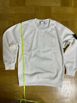 Vit sweatshirt från Stone Island  - Klassisk vit sweatshirt från stone island med rund halsringning och ribbade muddar vid ärmslut och nederkant. Tröjan har en patch på ena ärmen och är tillverkad i mjukt material, perfekt för en clean och enkel stil.