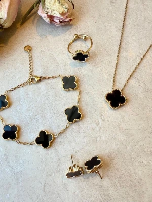 Set med svarta fyrklöver-smycken - Snyggt smyckeset med halsband, armband, ring och örhängen i guldfärgad metall. Alla delar har svarta fyrklöverformade hängen med guldfärgad kant. Perfekt för dig som gillar klassiska och trendiga accessoarer med unik design.
