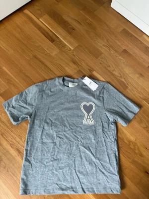 Ami Paris T-shirt  - Snygg grå t-shirt från Ami med ett stort hjärta och bokstaven A broderat på bröstet. Klassisk rund hals och korta ärmar. Tillverkad i mjuk bomull som känns skön mot huden. Perfekt för en clean och stilren look.