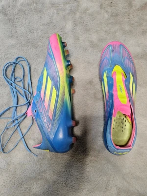Adidas F50 blåa fotbollsskor - Snygga Adidas F50 fotbollsskor i blått med neonrosa och gula detaljer. Skorna har klassisk låg profil, snörning och dobbar för spel på gräs. Tillverkade i syntetmaterial för lätt vikt och bra bollkontroll. Perfekta för dig som vill sticka ut på planen.
