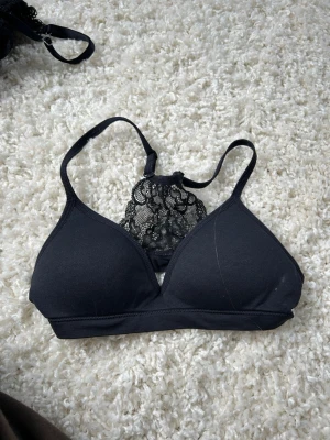 Svart bralette med spets från Lindex - Snygg svart bralette från Lindex med vadderade kupor och dekorativ spetsdetalj framtill. Axelbanden är smala och modellen är utan bygel för extra komfort. Tillverkad i en blandning med återvunnen polyamid. Perfekt som första bh då den är så sjukt bekväm!