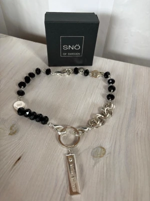 Halsband från SNÖ of Sweden med svarta pärlor - Snyggt halsband från SNÖ of Sweden med svarta, facetterade pärlor och silverfärgade länkar. Halsbandet har en rund ring och ett rektangulärt hänge med märkets logga. Perfekt för dig som gillar statement-smycken och vill sticka ut.