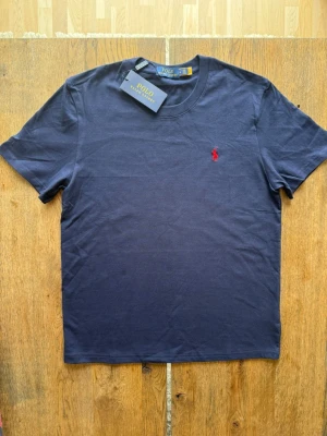 Mörkblå t-shirt från Polo Ralph Lauren - Snygg mörkblå t-shirt från Polo Ralph Lauren med klassisk röd logga broderad på bröstet. Tillverkad i mjuk bomull och har en normal passform med korta ärmar och rund hals. Perfekt basplagg för en stilren look.
