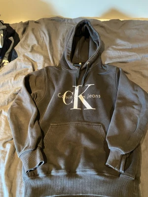 Svart hoodie från Calvin Klein Jeans - Säljer en svart hoodie från Calvin Klein Jeans med stor CK-logga och text på bröstet. Hoodien har huva med dragsko, magficka och ribbade muddar vid ärmar och nederkant. Perfekt för en avslappnad och trendig look.