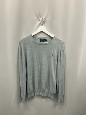 Grå rundhalsad tröja från Polo Ralph Lauren - Klassisk grå tröja från Polo Ralph Lauren med rund halsringning och diskret broderad logga på bröstet. Tröjan har långa ärmar och ribbade muddar vid ärmslut och nederkant. Perfekt basplagg i mjukt material.