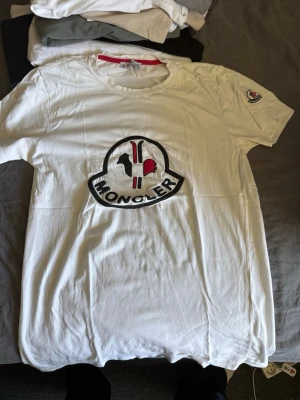 Vit Moncler t-shirt med stor logga - Säljer en vit t-shirt Moncler med stor broderad logga i svart, rött och vitt på bröstet. T-shirten har rund halsringning och en mindre Moncler-patch på ena ärmen. Tillverkad i mjukt bomullsmaterial och har en clean, klassisk passform.