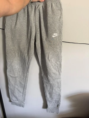 Gråa mjukisbyxor från Nike - Gråa mjukisbyxor från Nike med resår i midjan och muddar vid bensluten. Byxorna har en broderad vit Nike-logga på vänster ben och är gjorda i mjukt, fleeceliknande material på insidan. Perfekta för chill dagar eller träning.