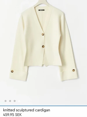 Kofta från gina - Superfin krämvit stickad cardigan från Ginatricot med skulpterad form och stora bruna knappar framtill och vid ärmsluten. V-ringad och croppad modell med långa ärmar. Perfekt att styla med jeans eller kjol. Har fläckar vid slutet av ärmarna från smink.