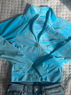 Blå velourtröja från Juicy Couture, Strl XS - Säljer en ikonisk blå velourtröja från Juicy Couture med dragkedja och hög krage. Tröjan har långa ärmar, ribbade muddar och glittrande Juicy-text på ryggen. Perfekt för dig som gillar Y2K-stil och vill ha något mjukt och bekvämt.