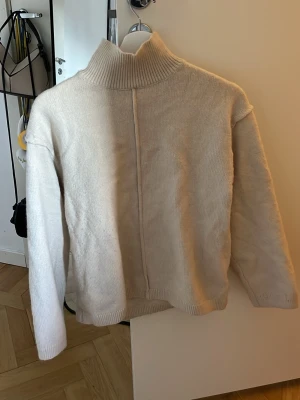 Beige polotröja från H&M - Säljer en beige stickad polotröja från H&M i storlek XS. Tröjan har hög krage, lång ärm och en synlig söm framtill som detalj. Perfekt för kalla dagar och enkel att matcha med olika outfits.
