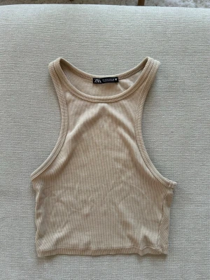 Beige Croptop -  beige croptop från Zara med ribbad struktur. Toppen är ärmlös och har en sportig, tight passform med hög halsringning. Perfekt att styla med jeans eller kjol för en trendig look. Inte använd på flera år 