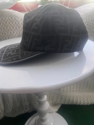 Svart keps med Fendi-logga - Snygg svart keps med Fendis ikoniska FF-mönster över hela kepsen. Klassisk böjd skärm och justerbar passform. Tillverkad i tyg med exklusiv känsla och passar perfekt för dig som vill ha en trendig accessoar med lyxig touch.