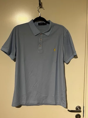 Ljusblå pikétröja från Polo Ralph Lauren - Säljer en ljusblå pikétröja från Polo Ralph Lauren med klassisk krage och tre knappar framtill. Tröjan har korta ärmar och den ikoniska gula broderade loggan på bröstet. Perfekt till jeans eller shorts för en clean look.