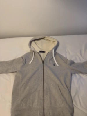 Grå hoodie från Polo Ralph Lauren - Snygg grå hoodie från Polo Ralph Lauren med dragkedja och vit snörning i huvan. Klassisk logga broderad på bröstet och mjukt bomullsmaterial. Perfekt för en chill och stilren look.