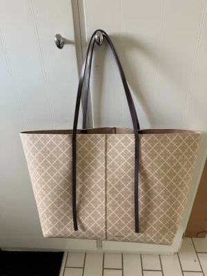 Beige shoppingväska By Malene Birger - Stilren shoppingväska i beige med ljusbrunt grafiskt mönster från By Malene Birger. Väskan har långa, smala axelband i mörkbrunt och är tillverkad i ett tåligt konstlädermaterial. Perfekt för dig som vill ha en rymlig och trendig väska.