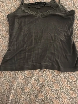 Mörkgrön linnetopp med spets - Snygg mörkgrön linnetopp från The Black Label Collection i storlek M. Toppen har tunna axelband och dekorativ spets längs den v-ringade urringningen. Perfekt att styla med jeans eller kjol för en trendig look.