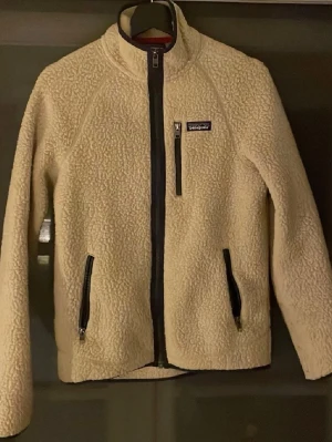 Beige fleecejacka från Patagonia - Beige fleecejacka från Patagonia med hög krage och dragkedja framtill. Jackan har tre fickor med dragkedja och en liten logga på bröstet. Perfekt nu till hösten.
