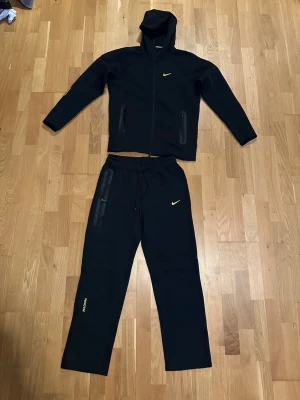 Svart Nike loungewear set med huva - Snyggt svart loungewear set från Nike med collab med Nocta. Storlek M men passar bra för S med. Skriv för mer bilder eller frågor allmänt. Har använt lite sedan bilderna tagits men inga stora skavanker trycket på några få detaljer har börjat lossna