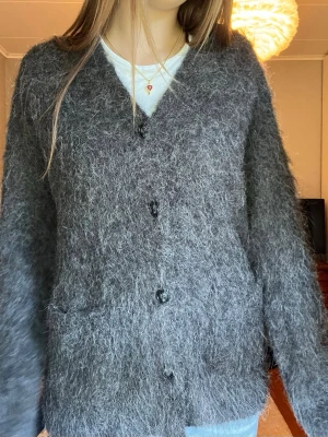 Mohair kofta med knappar - Mysig grå mohair kofta och knäppning framtill. Koftan har v-ringning, långa ärmar och två fickor framtill. Perfekt för lager-på-lager och en chill look. Aldrig använd