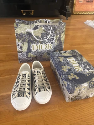 Dior sneakers med monogrammönster - Snygga låga sneakers från Dior med klassiskt monogrammönster i blått och vitt. Skorna har vita snören, vit sula och detaljer med texten 'Christian Dior' på plösen och sidan. Perfekta för dig som vill ha en exklusiv och trendig look.