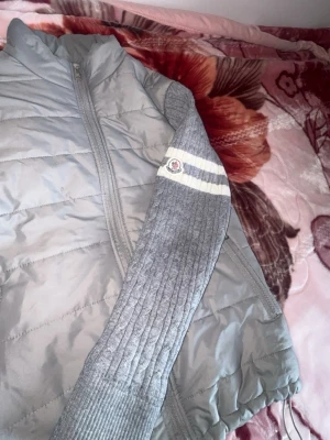 Grå quiltad cardigan med stickade ärmar - Snygg grå cardiganjacka med quiltad kropp och stickade ärmar. Jackan har dragkedja framtill, två fickor med dragkedja och vita ränder på ärmarna. Perfekt för lager-på-lager och chill still sätt kan diskuteras