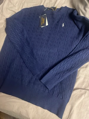 Blå kabelstickad tröja Polo Ralph Lauren - Snygg blå kabelstickad tröja från Polo Ralph Lauren med rund halsringning och den klassiska broderade loggan på bröstet. Tröjan har långa ärmar och ribbade muddar vid ärmslut och nederkant. Perfekt för dig som gillar stilren och tidlös design. Sitter som M