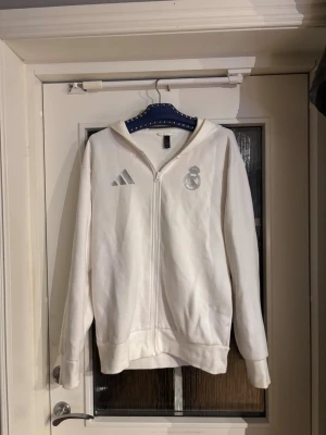 Vit Real Madrid hoodie med dragkedja - Säljer en vit hoodie från Adidas med Real Madrid-logga på bröstet och texten 'REAL MADRID' på ryggen. Hoodien har dragkedja framtill, två fickor och en klassisk huva. Perfekt för dig som älskar fotboll och vill ha en clean och sportig stil.